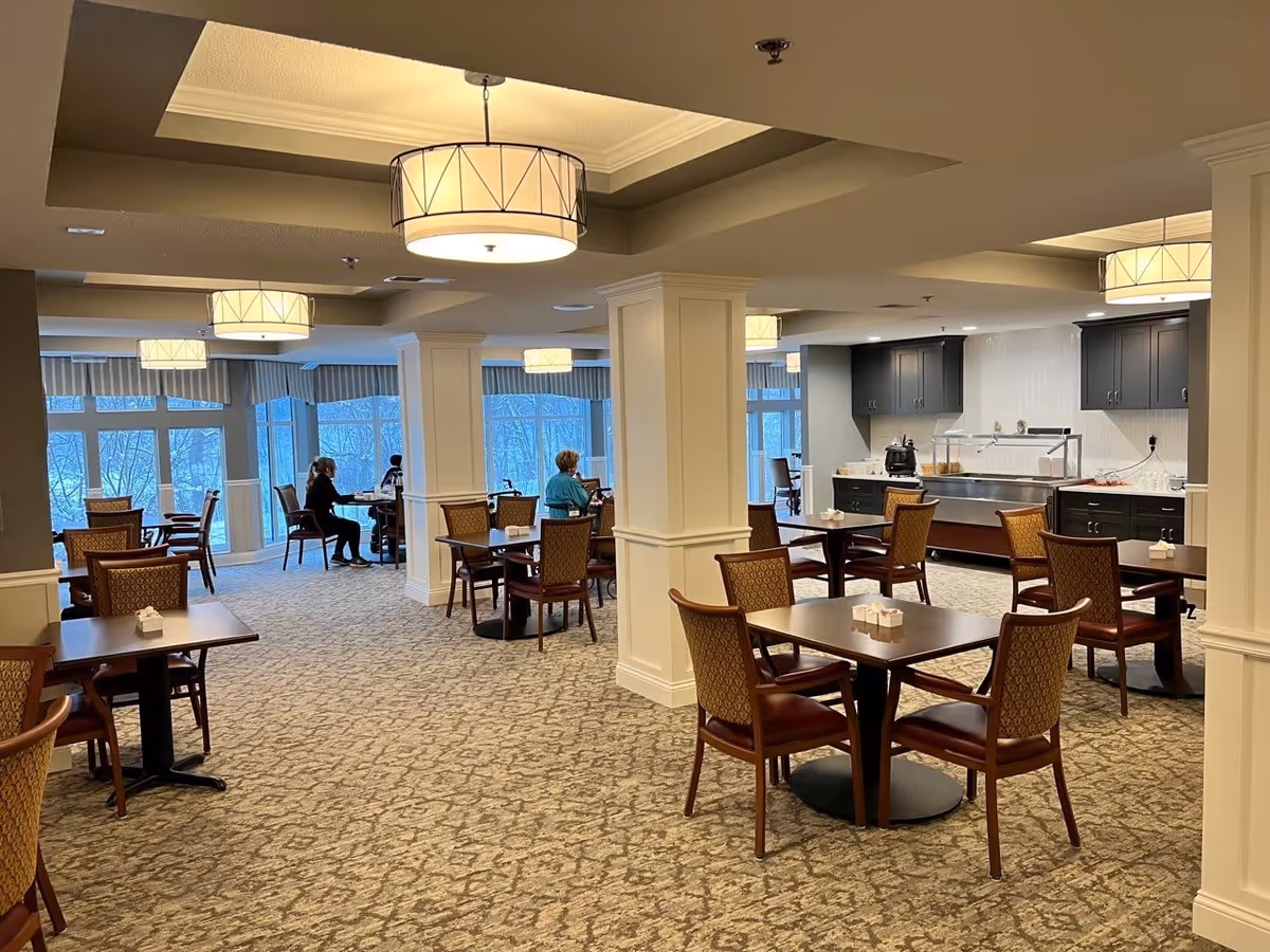 A spacious dining area in a senior living facility with several tables and chairs arranged neatly. There are a few elderly individuals seated at some tables near large windows with a view of trees outside. The room has beige carpet, white pillars, and modern ceiling lights. A serving area with cabinets and a soup pot is visible in the background.