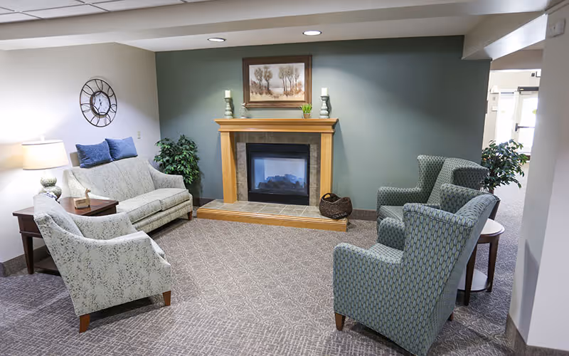 A cozy senior living facility common area with a fireplace centered on a green accent wall. The room features a light-colored sofa with blue cushions, two patterned armchairs, a side table with a lamp, and a wall clock. There are decorative plants and a framed painting above the fireplace.