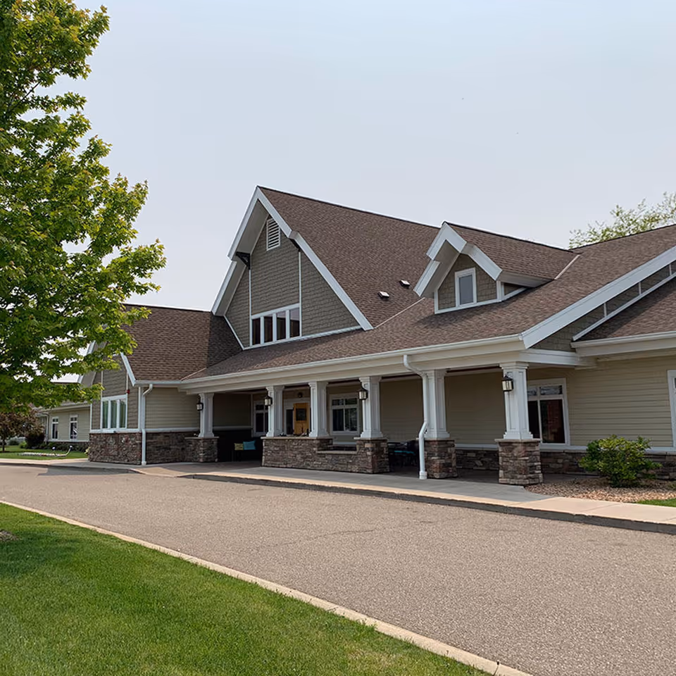 Exterior view of a single-story assisted living facility building with a covered entrance supported by white columns with stone bases, a brown shingled roof, beige siding, and a tree with green leaves on the left side.