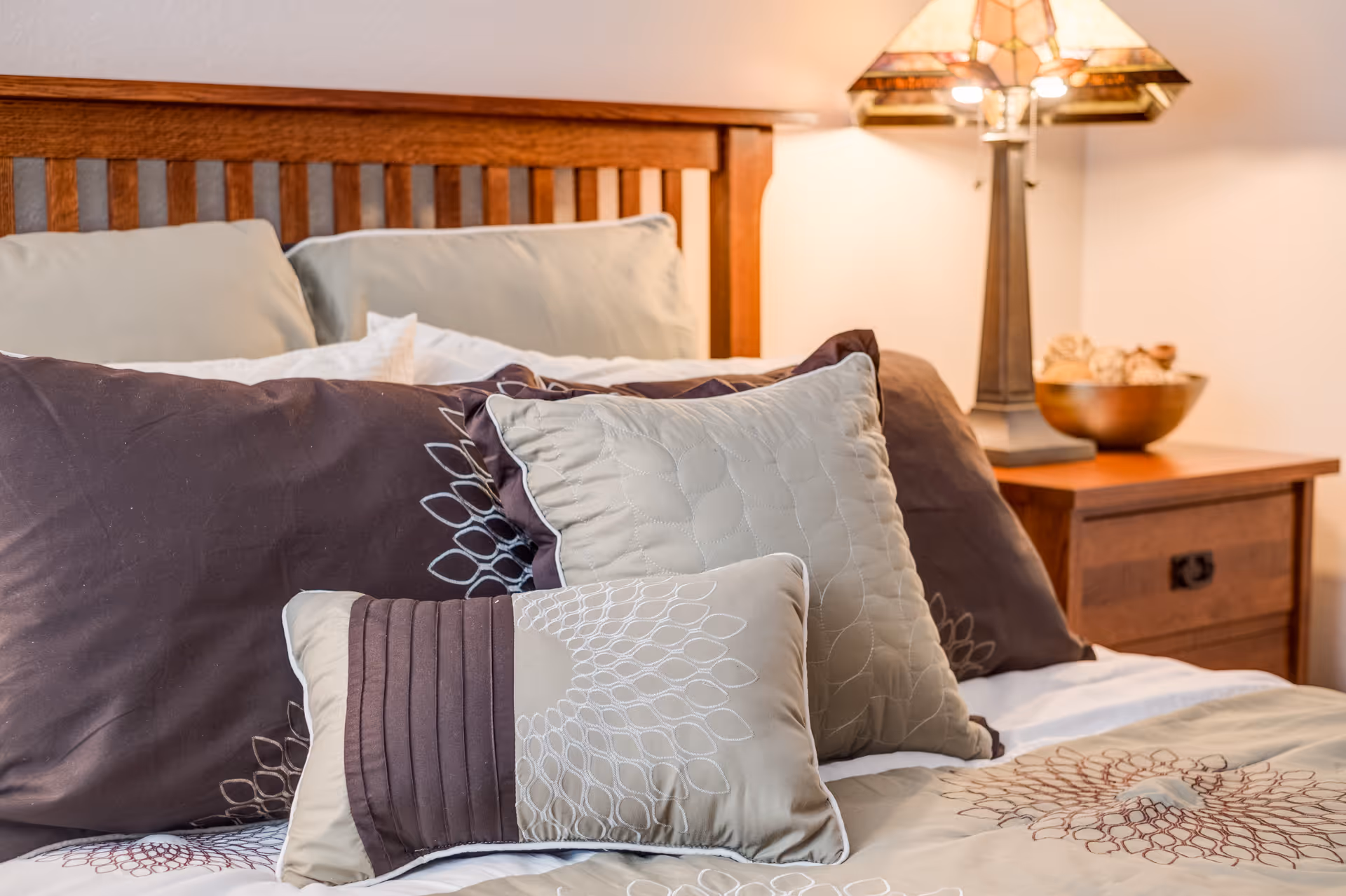 Close-up of a neatly made bed with decorative pillows, a wooden headboard, and a bedside table with a lamp.