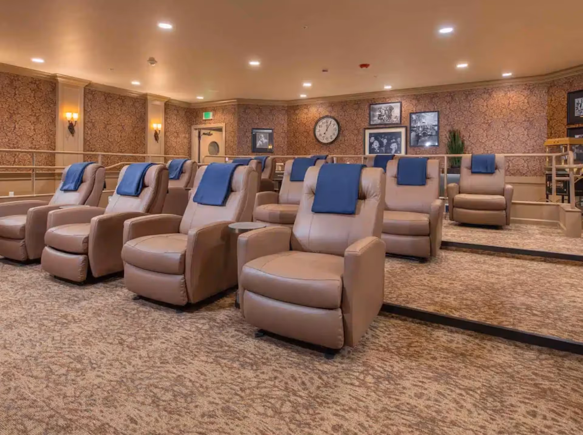 Interior theater-style lounge with rows of beige recliners draped with blue cloths, carpeted floor, and framed pictures on patterned walls.