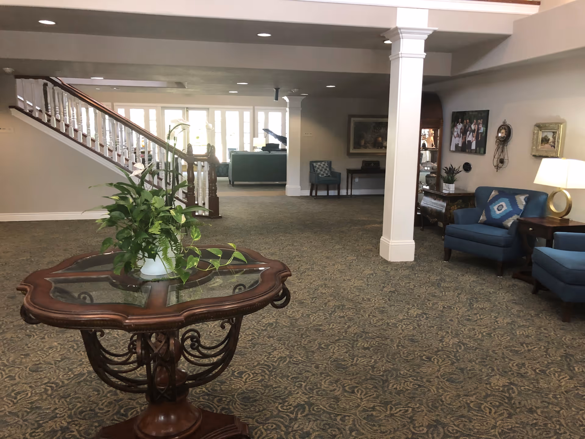 Spacious interior common area with patterned carpet, a wooden table with a potted plant in the foreground, blue upholstered chairs with decorative pillows, a lamp on a side table, framed pictures on the wall, and a staircase leading to an upper floor.