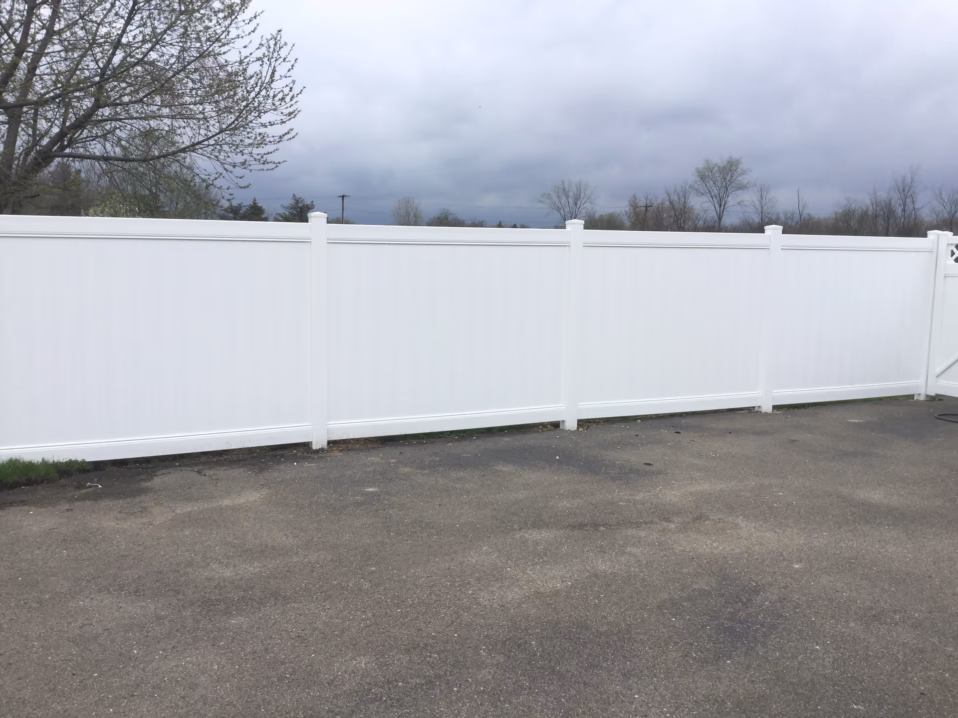 A long white vinyl privacy fence with vertical panels and posts, set on a paved surface with some grass visible at the base. In the background, there are leafless trees and a cloudy sky.