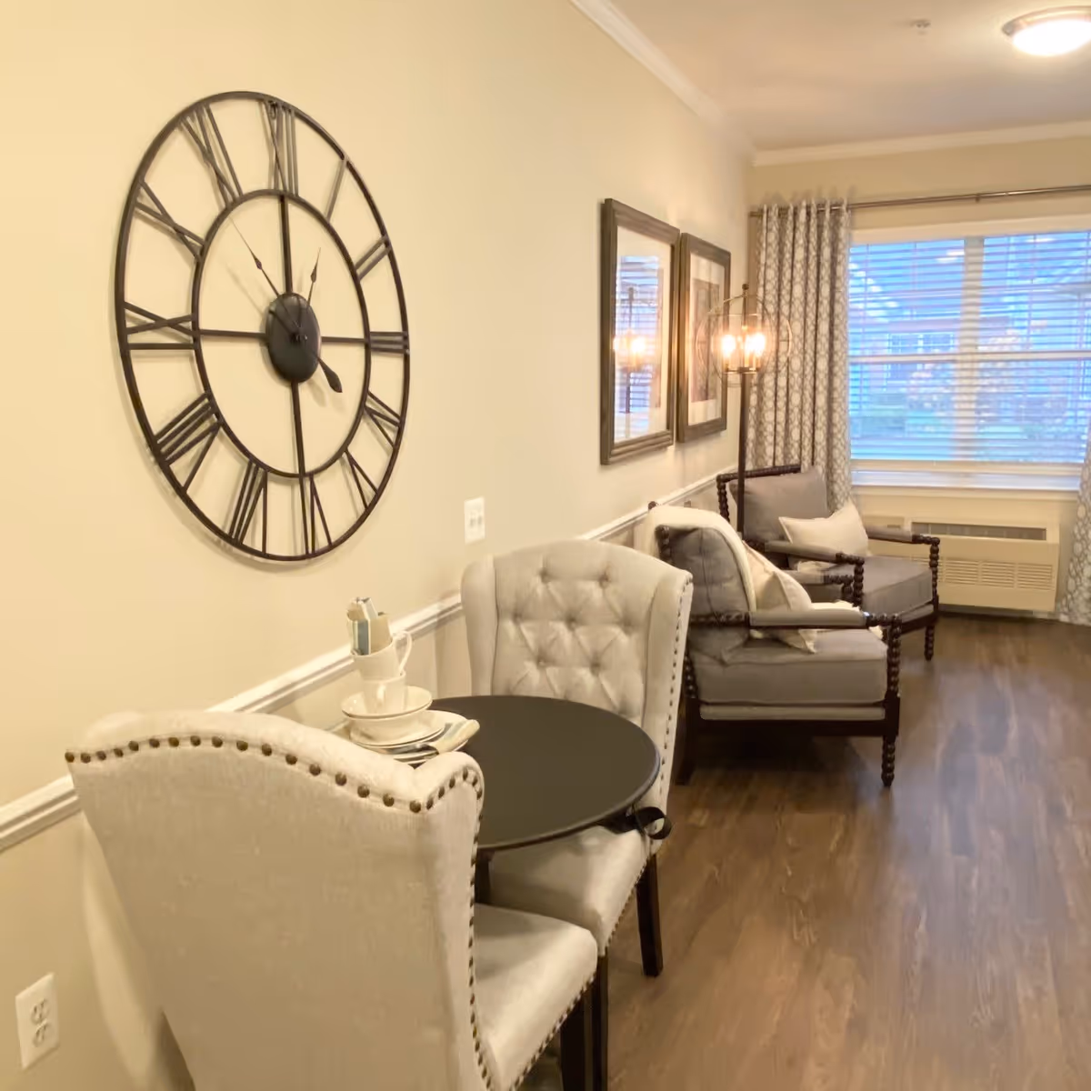 A cozy living room area with two upholstered chairs around a small round black table set with cups and plates. On the wall above the table is a large decorative clock with Roman numerals. Further into the room are two cushioned armchairs with pillows, a floor lamp with exposed bulbs, two framed pictures on the wall, and a window with patterned curtains letting in natural light. The floor is wooden.