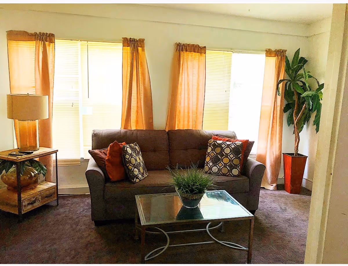 A cozy living room with a brown sofa adorned with patterned and solid orange cushions, a glass coffee table with a small green plant, a side table with a lamp and decorative items, orange curtains on three windows, and a tall potted plant in a red planter in the corner.