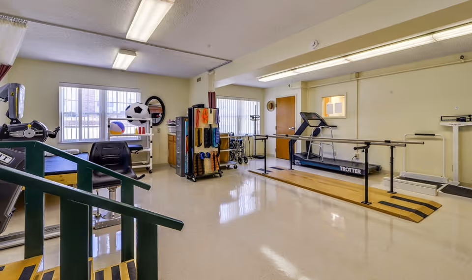 A rehabilitation or physical therapy room with exercise equipment including a treadmill, parallel bars for walking practice, a stationary bike, therapy balls, resistance bands, and a set of stairs for physical therapy exercises. The room has large windows with vertical blinds, fluorescent lighting, and a clean, polished floor.