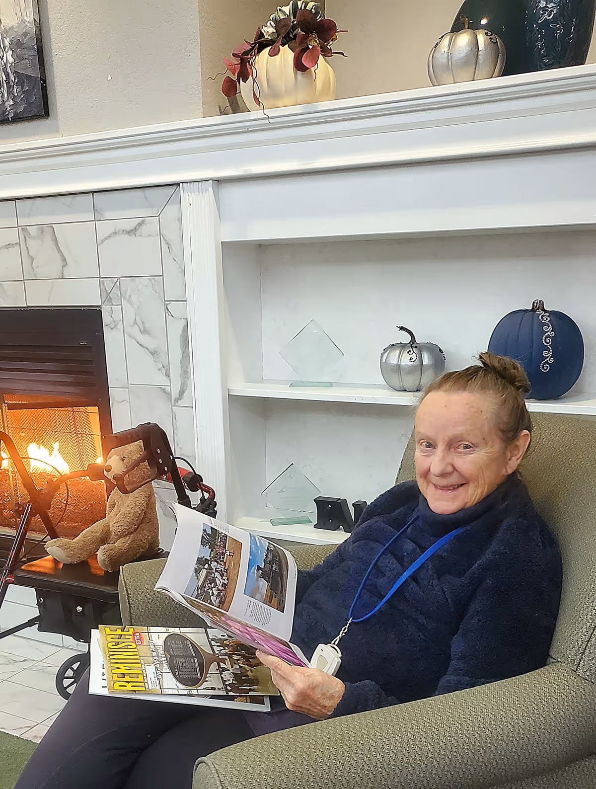 An elderly woman sitting comfortably in a green armchair by a fireplace with a lit fire. She is smiling and holding an open magazine, with another magazine resting on her lap. Behind her is a white built-in shelf decorated with small pumpkins and glass awards. A teddy bear is sitting on a walker near the fireplace.