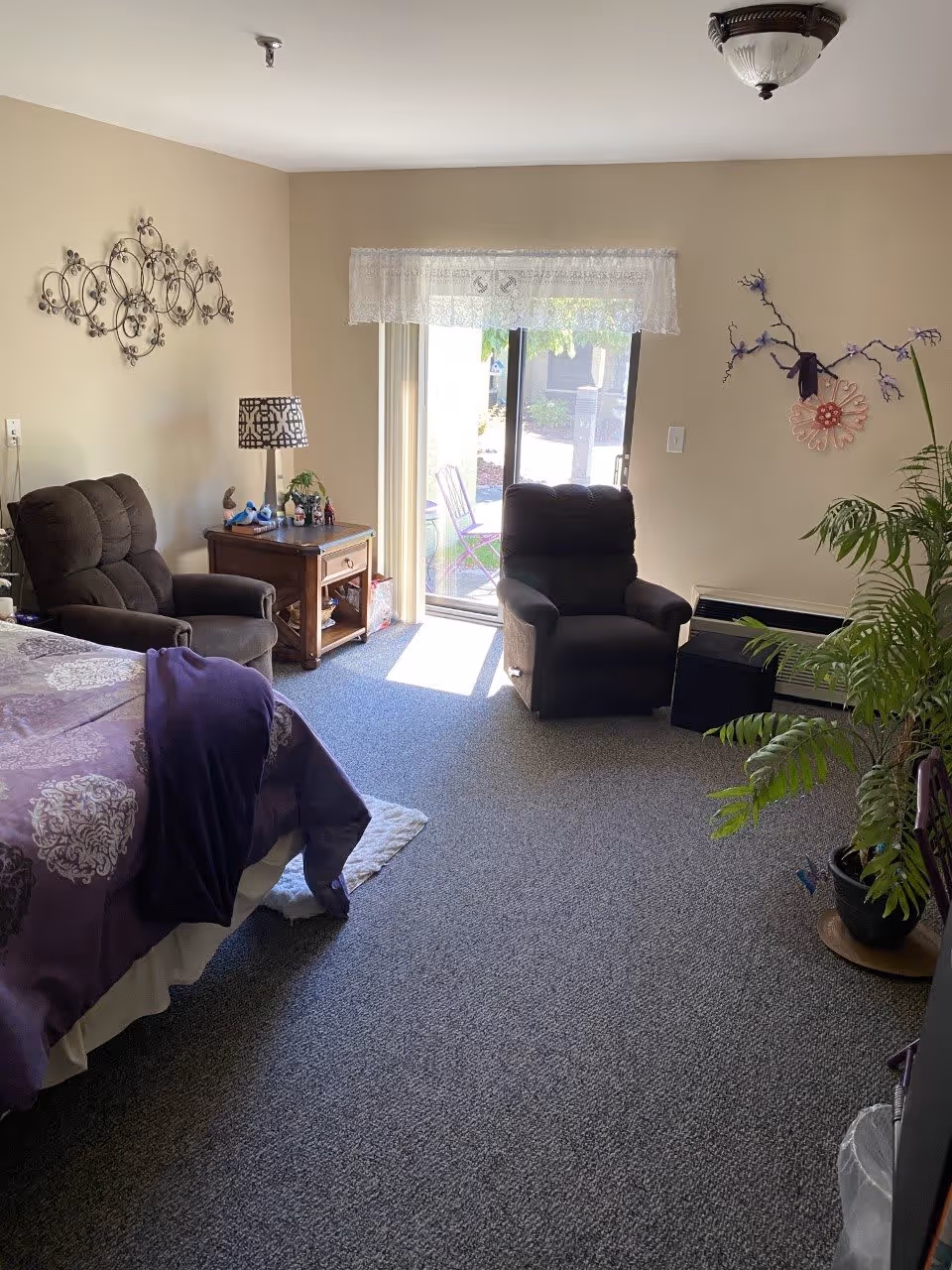 A cozy bedroom with a bed covered in purple bedding, two dark armchairs, a wooden nightstand with a lamp and small decorations, a sliding glass door with sheer white curtains leading outside, and a potted plant near the wall with decorative wall art.