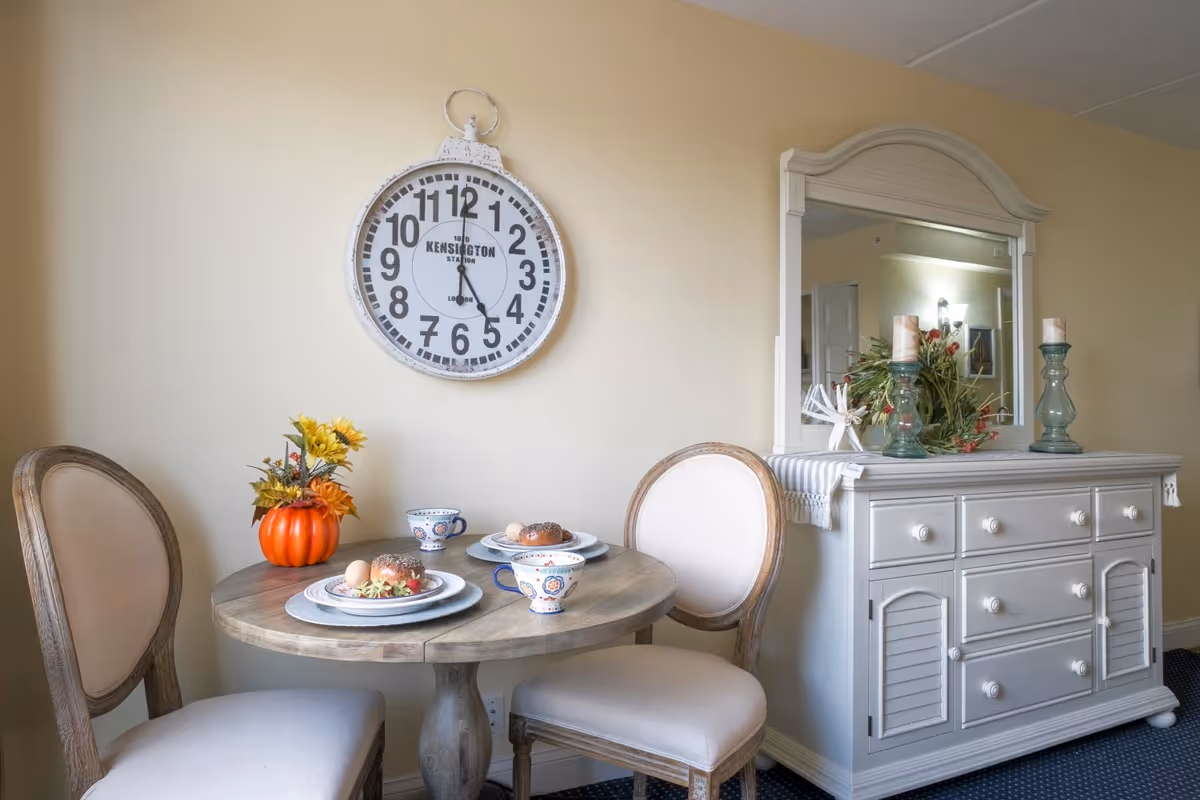 A small round wooden dining table set for two with plates of food, cups, and a small pumpkin centerpiece with flowers. Two upholstered chairs are placed around the table. On the wall above the table is a large round clock. To the right is a white dresser with a mirror, decorated with candles and a floral arrangement.