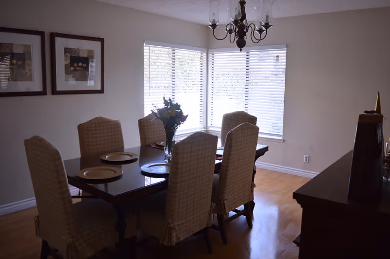 Dining room with a rectangular table surrounded by six upholstered chairs, a vase of flowers in the center, and a bright bay window with blinds.