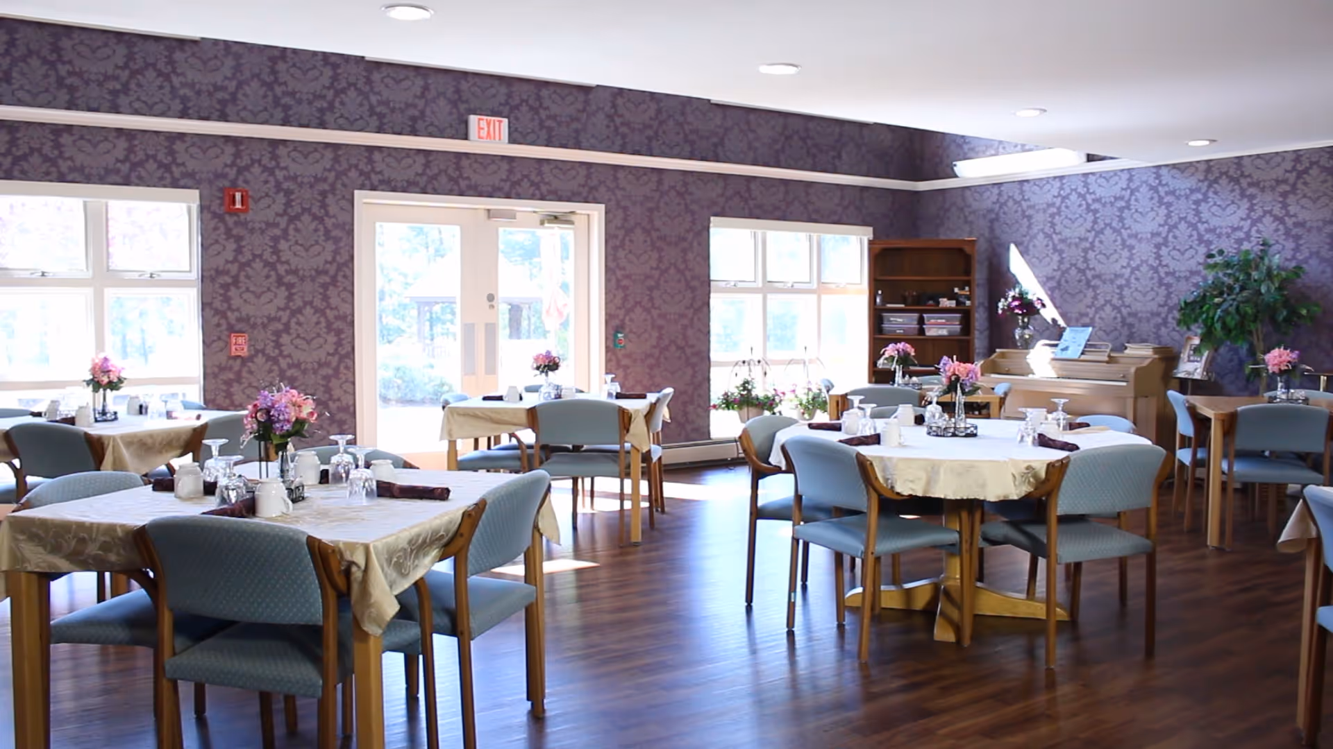 Dining room with tables set with tablecloths, floral centerpieces, upholstered chairs, and patterned purple wallpaper.