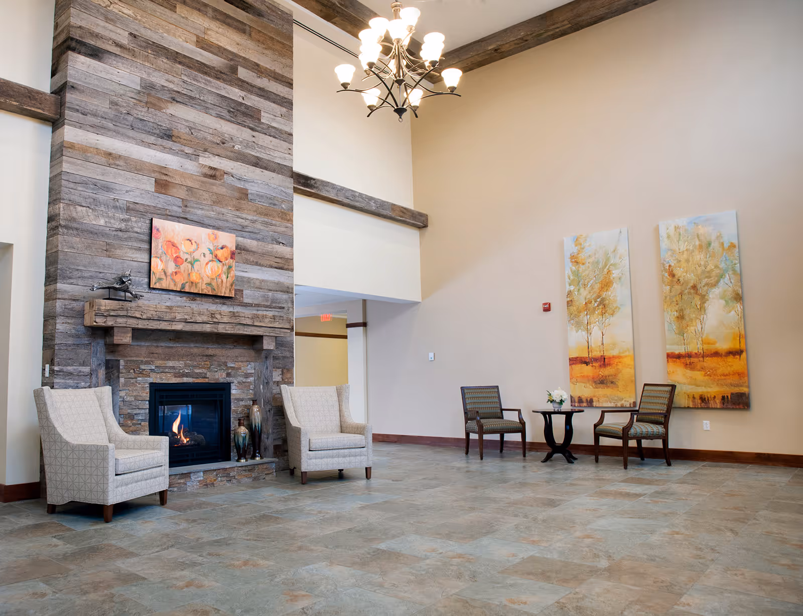 A spacious living area with a tall wooden fireplace surrounded by stone, two beige armchairs placed on either side of the fireplace, and a chandelier hanging from the ceiling. On the right side, there are two wooden chairs with patterned cushions and a small round table with a flower arrangement. Two large paintings of trees with autumn colors hang on the wall behind the chairs.