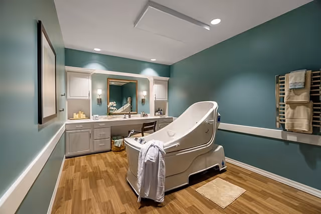 A spacious bathroom with teal walls and wood flooring featuring a large white therapeutic bathtub with a white robe hanging on its side. The room includes a vanity with a large mirror, two wall sconces, and various toiletries. Towels are neatly arranged on a wall-mounted rack.