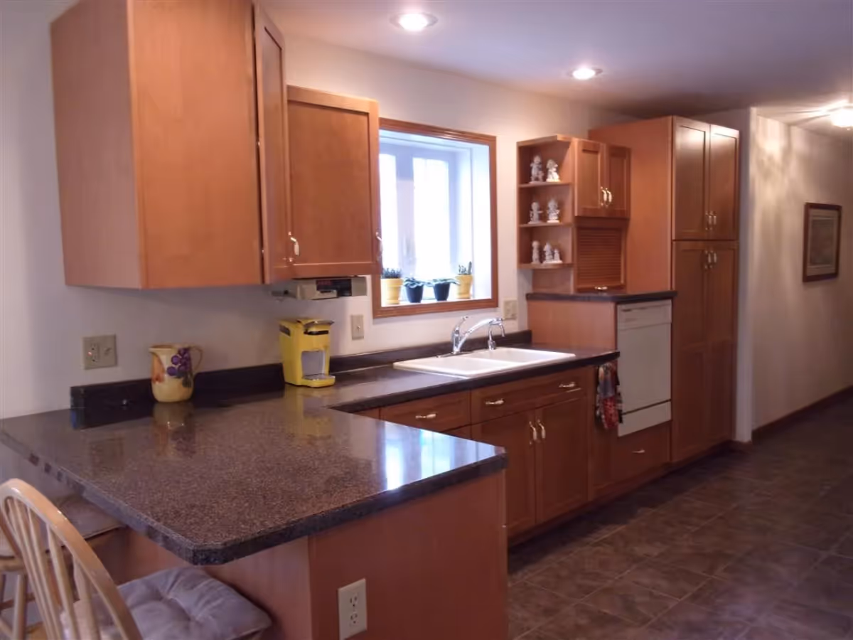 Interior view of a kitchen with wooden cabinets, a double sink under a window with potted plants, a yellow coffee maker on the counter, and a tiled floor. There are two cushioned wooden chairs at the counter and recessed ceiling lights.