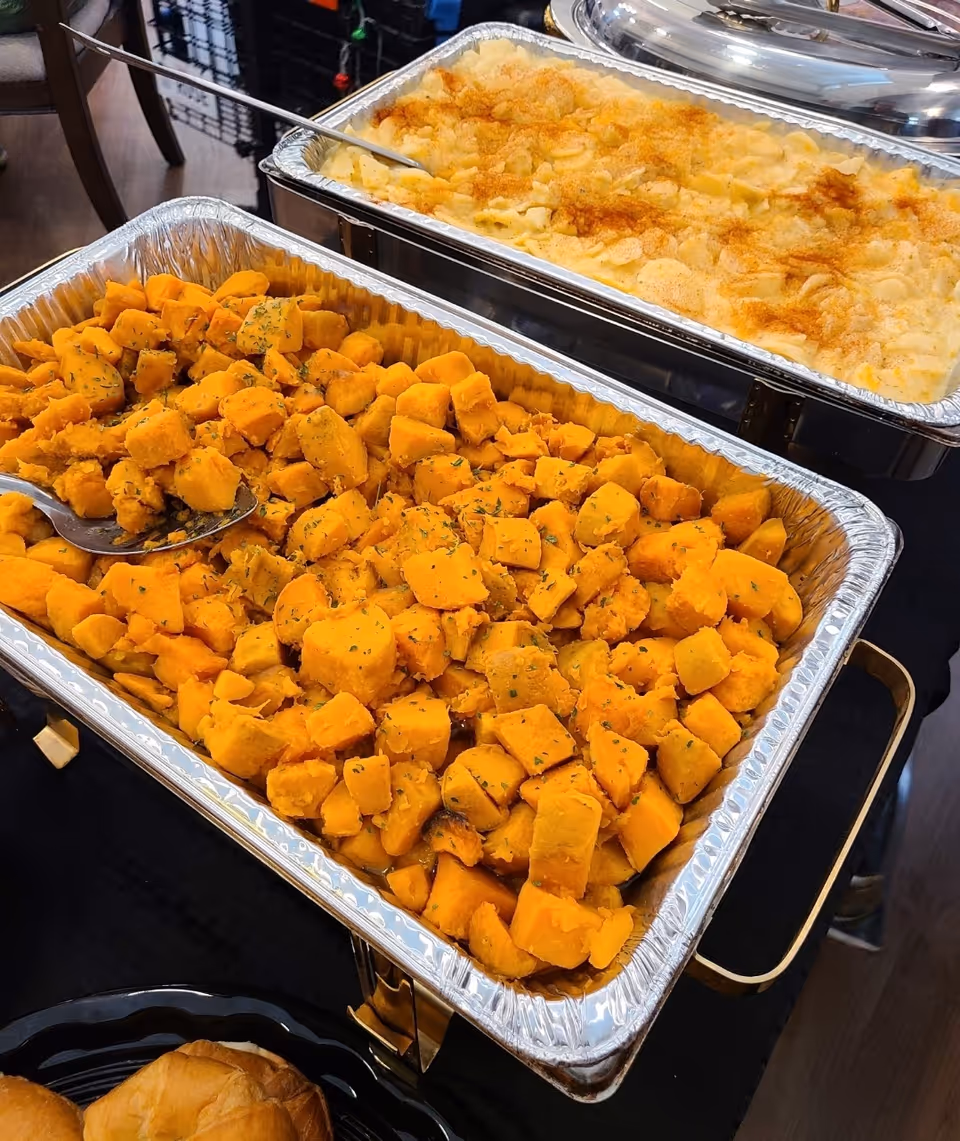 Two large aluminum trays filled with cooked food on a buffet table. The front tray contains seasoned cubed sweet potatoes, and the back tray contains macaroni and cheese topped with a golden breadcrumb crust. A serving spoon is placed in the sweet potatoes tray.