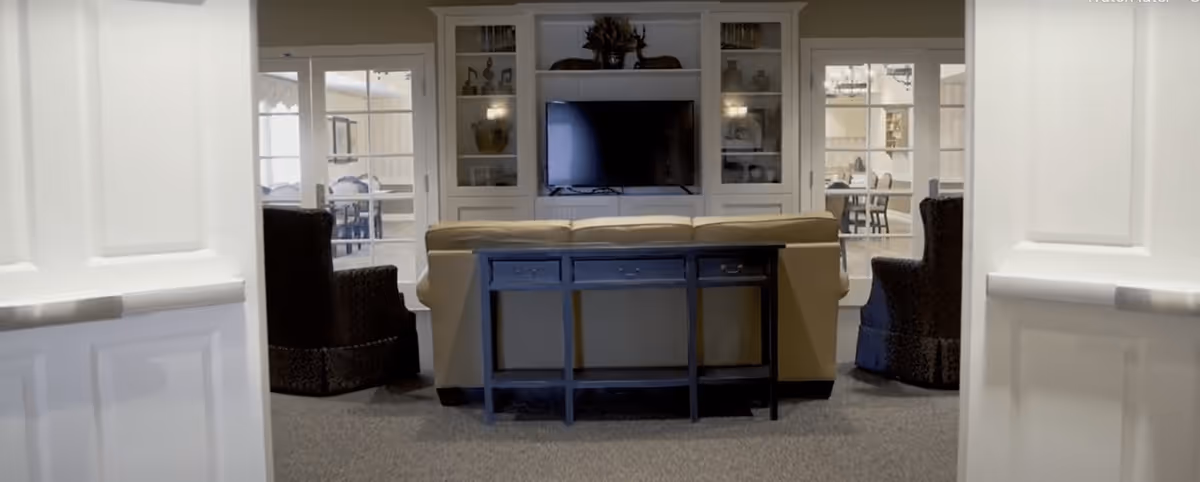 View through double doors into a cozy living room area with a beige sofa facing a flat-screen TV mounted on a white built-in shelving unit. Two dark upholstered armchairs flank the sofa. Beyond the living room, a dining area with tables and chairs is visible through glass doors.