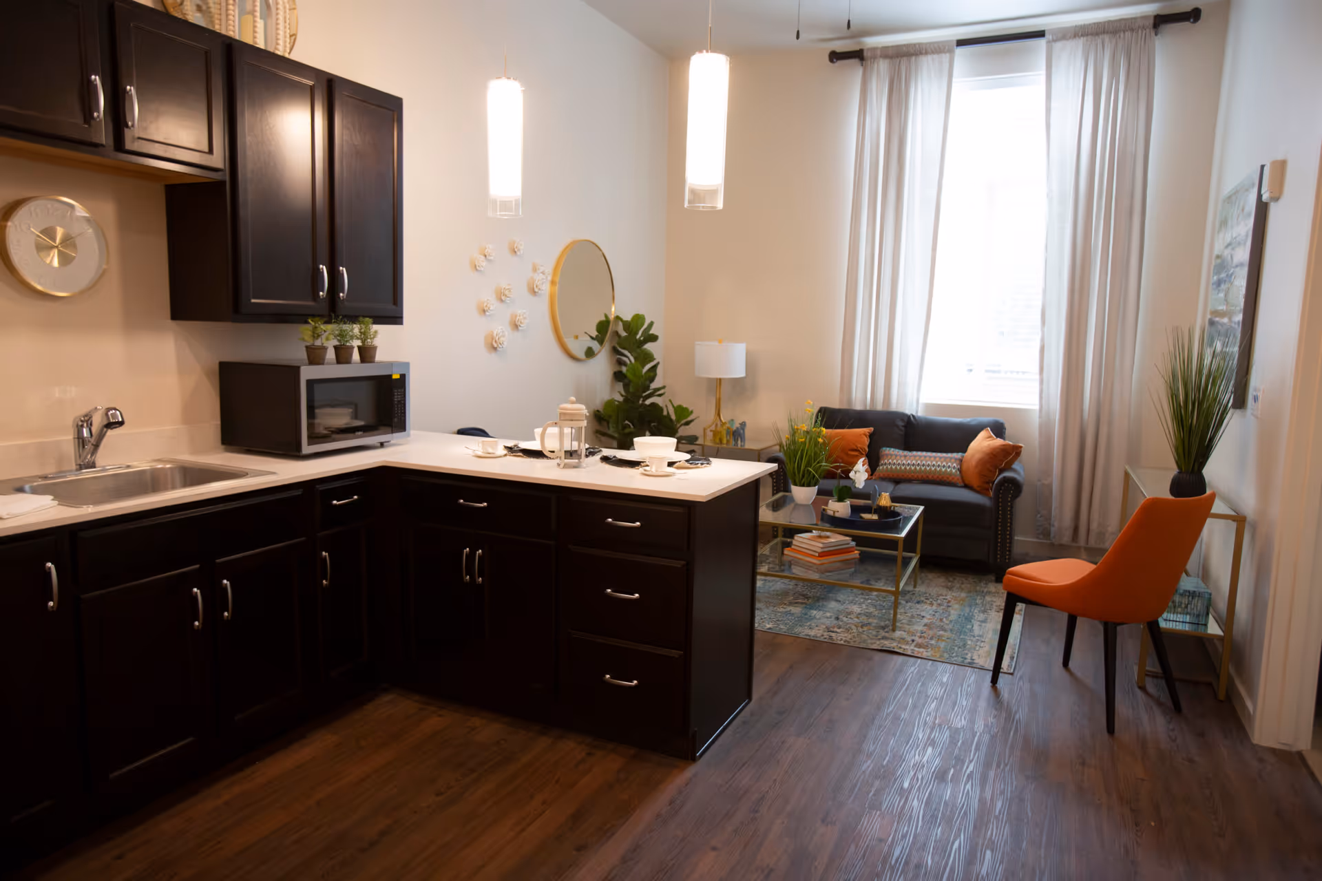 Open-plan interior showing a dark wood kitchen with a white island opening into a living area with a sofa, orange chair, coffee table and a large window.