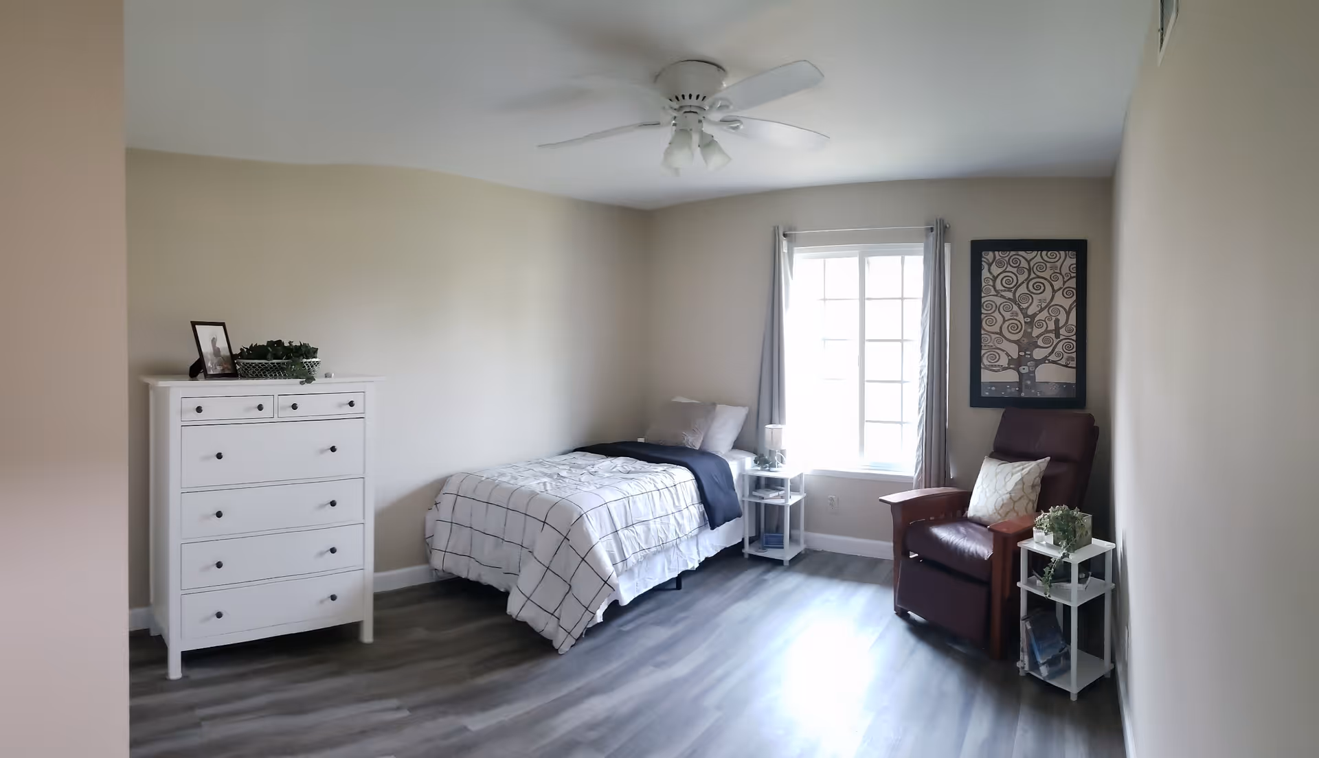 A simple, clean bedroom with a single bed covered in a white and black checkered blanket, a white dresser with a framed photo and a plant on top, a window with gray curtains, a brown armchair with a white pillow, two small white side tables with plants and books, and a ceiling fan with lights.