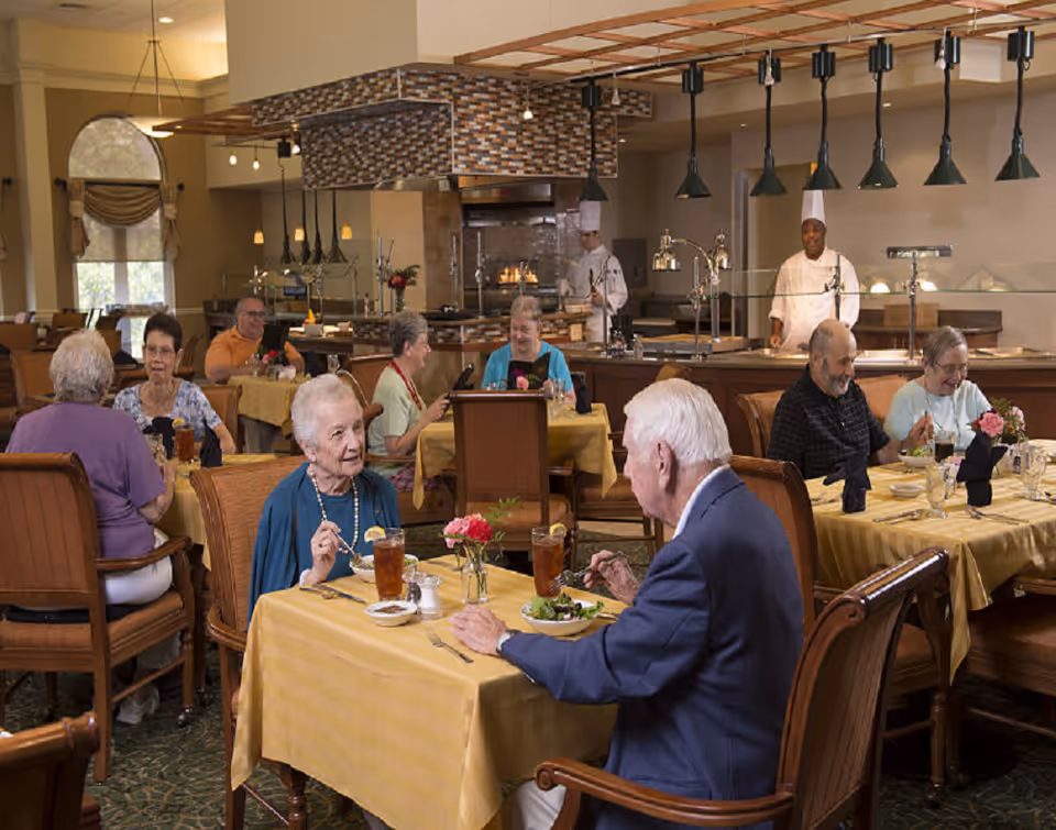 Several elderly residents dine and converse at tables in a well-lit senior living dining room with chefs working behind a serving counter.