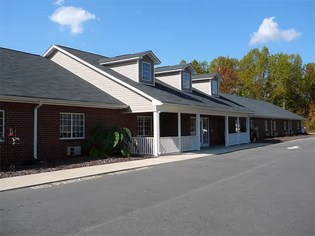 Exterior view of a single-story brick building with a gray shingled roof and white trim. The building has a covered porch with white railings and three dormer windows on the roof. There is a paved driveway in front and trees with autumn foliage in the background under a partly cloudy sky.