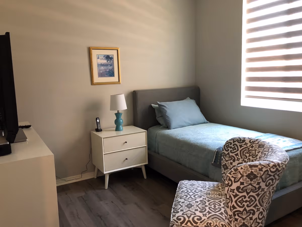 A small bedroom with a single bed made with light blue bedding and pillows. Next to the bed is a white nightstand with two drawers, a turquoise lamp, and a cordless phone. A patterned armchair is positioned near the foot of the bed. A framed picture hangs on the beige wall above the nightstand, and a window with striped blinds lets in natural light.