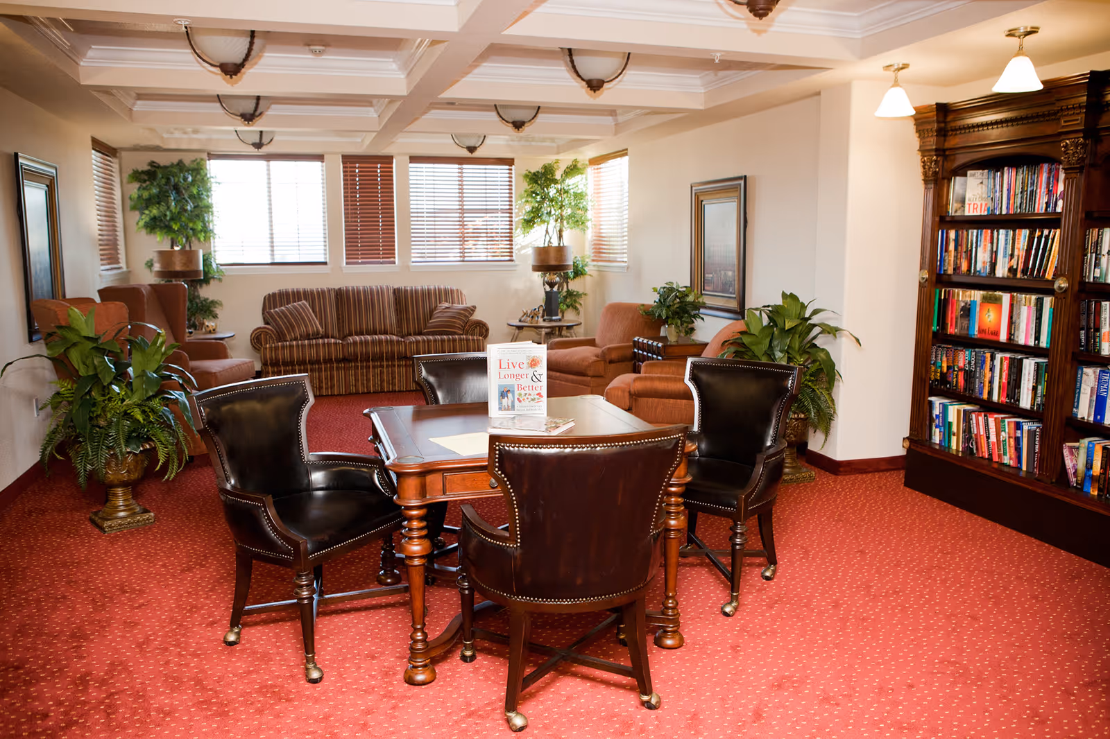 A cozy common area in Meadowlark Estates Gracious Retirement Living featuring a wooden table with four leather chairs on a red carpet. In the background, there are two striped sofas, two armchairs, several potted plants, a wooden bookshelf filled with books, and windows with blinds letting in natural light.