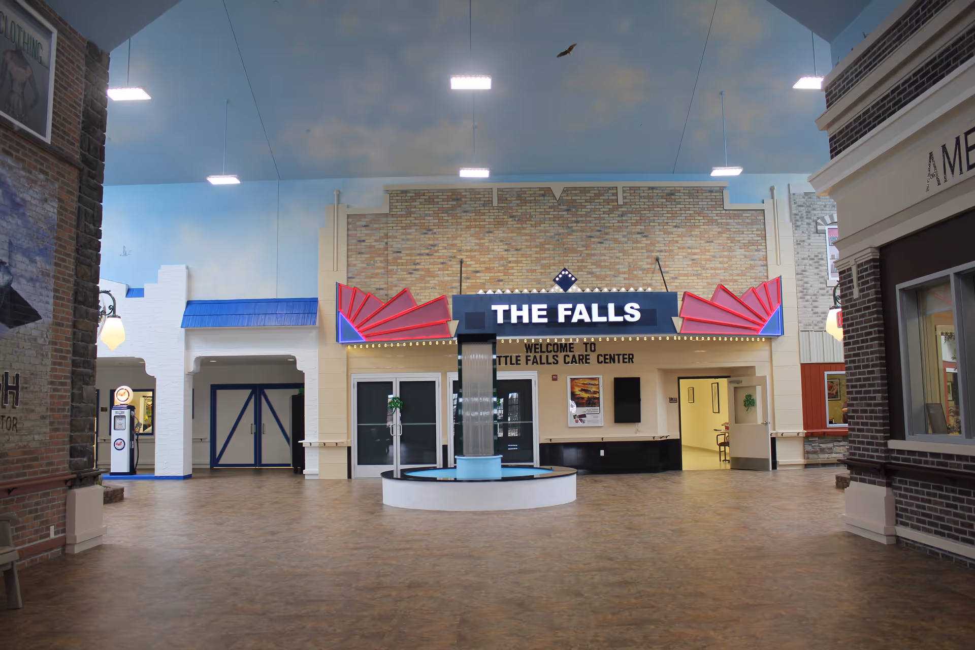 Interior view of a senior living facility lobby area with a decorative sign reading 'THE FALLS' above an entrance labeled 'WELCOME TO LITTLE FALLS CARE CENTER'. The space has a high ceiling painted with a sky and clouds motif, a central water fountain, and various storefront-style facades along the walls.