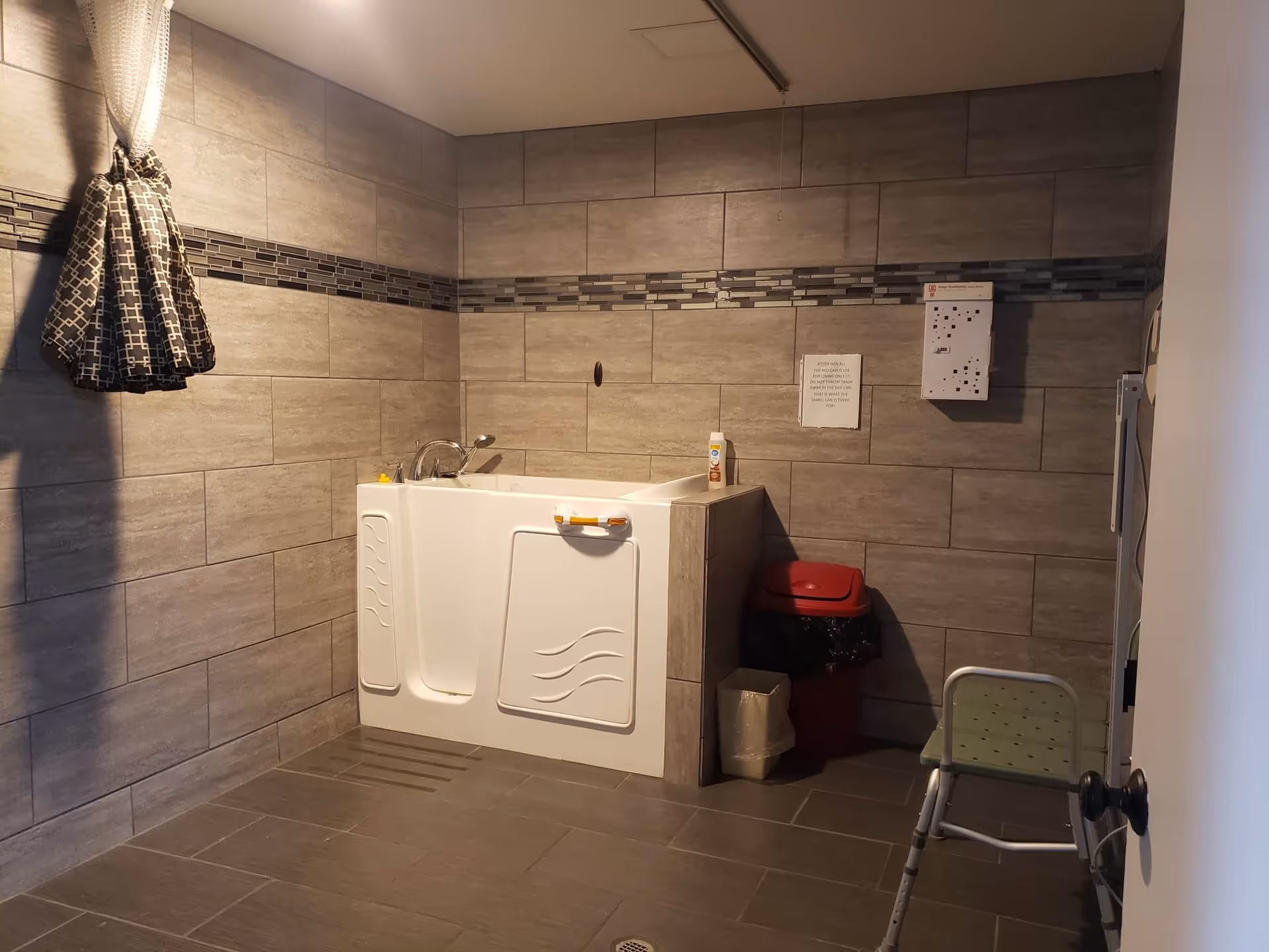 A bathroom with gray tiled walls and floor featuring a white walk-in bathtub with safety handles. There is a shower chair in the corner, a red trash bin with a black liner, a small beige trash bin, and a hanging patterned shower curtain on the left side. A bottle of lotion or soap is placed on the bathtub ledge, and there are two signs posted on the wall above the trash bins.