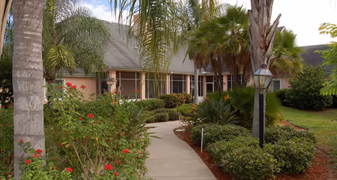 Front entrance of a single-story building with a curved walkway, palm trees, and landscaped gardens.