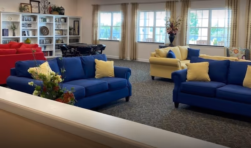 A spacious living room area with blue and yellow sofas adorned with yellow and blue pillows. There is a red sofa in the background near a white bookshelf filled with books and decorative items. The room has large windows with beige curtains allowing natural light to fill the space. A dining table with chairs is also visible near the bookshelf.