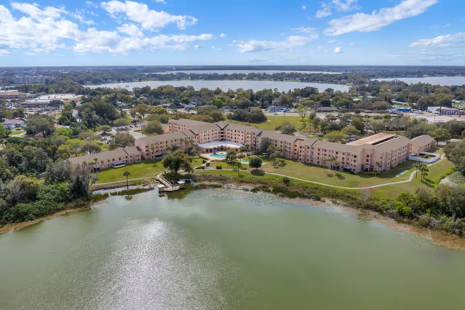 Aerial view of a large senior living facility named Spring Haven situated next to a body of water with a dock. The building is surrounded by green lawns, trees, and walking paths, with a swimming pool visible in the courtyard. The background shows a suburban area with more trees and buildings under a partly cloudy sky.