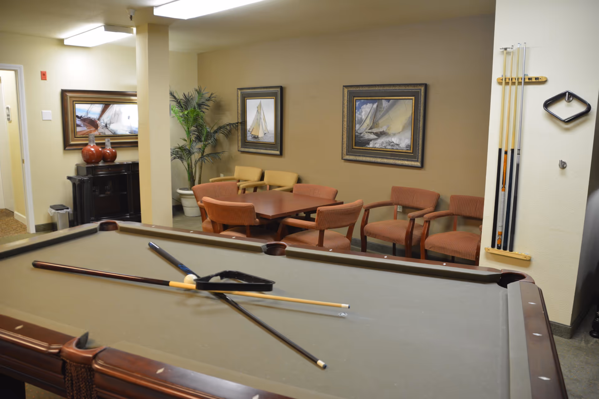 Interior view of a recreational room featuring a pool table with cues and a triangle rack on it. Behind the pool table, there is a seating area with a square wooden table surrounded by six upholstered chairs in red and yellow tones. The walls are decorated with framed pictures of sailboats, and there is a potted plant in the corner. A rack holding pool cues is mounted on the wall to the right.