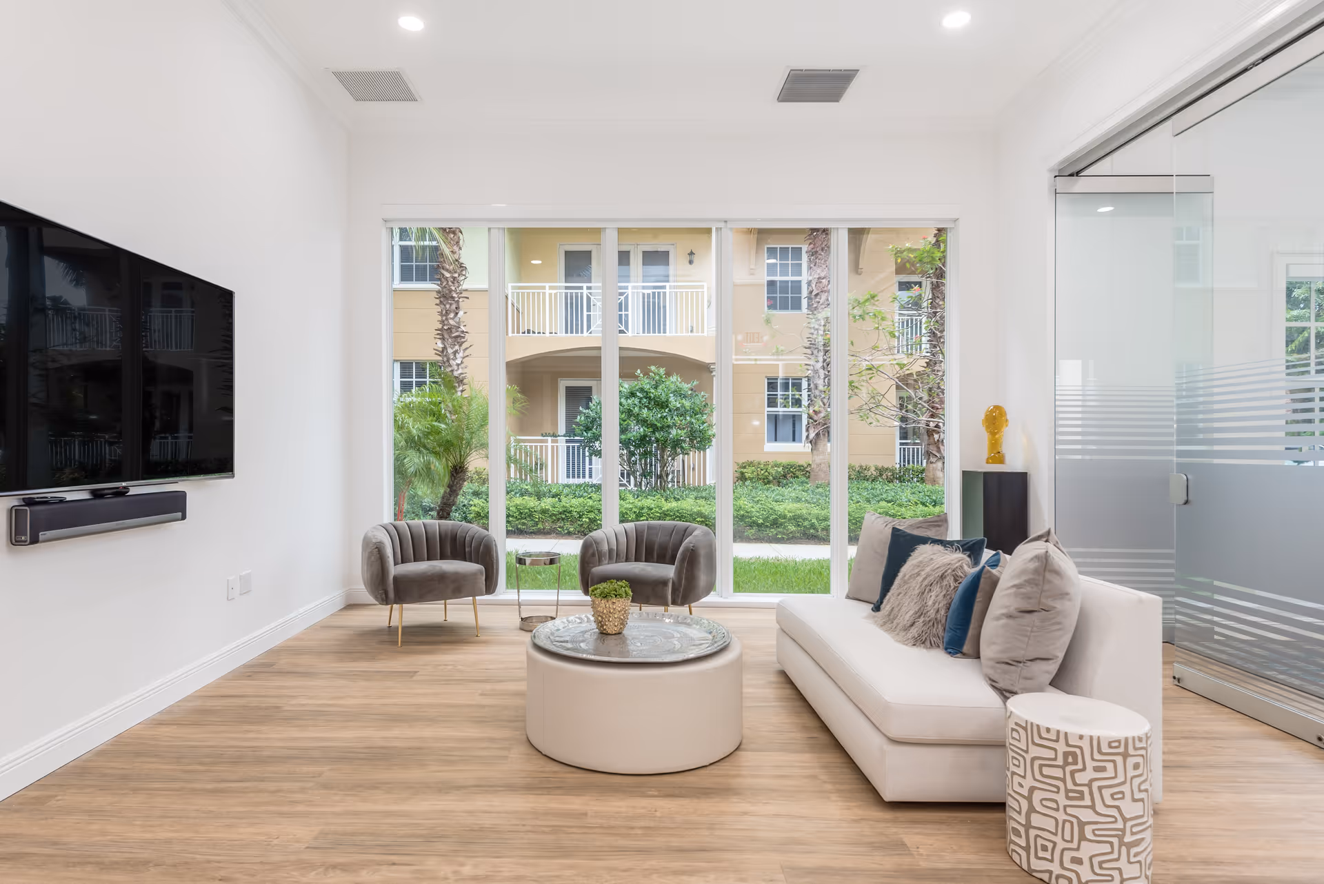 A modern living room with a white sofa adorned with multiple cushions, two gray armchairs, a round coffee table with a small plant on it, and a large flat-screen TV mounted on the wall. The room has large floor-to-ceiling windows showing a view of greenery and an adjacent building. The floor is wooden, and there is a glass door on the right side.
