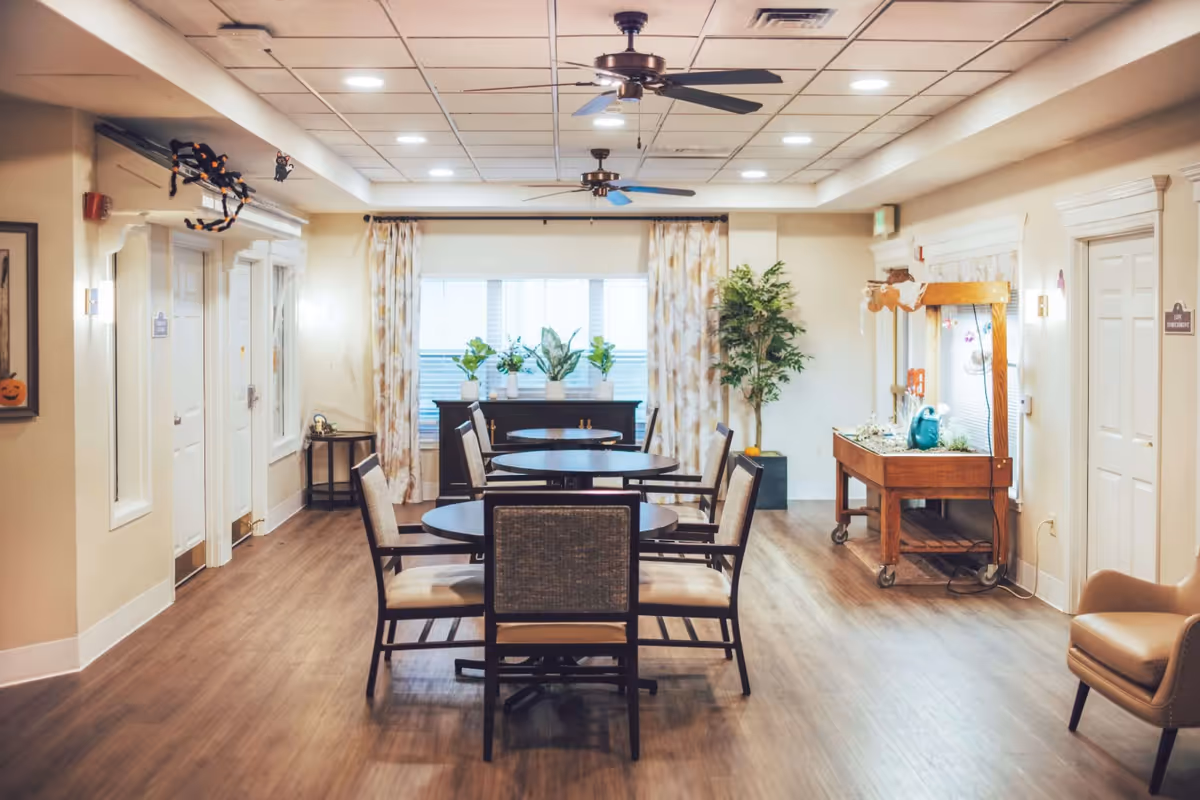 A well-lit interior room with round tables and chairs arranged for seating. The room has wood flooring, ceiling fans, and large windows with patterned curtains. There are plants on a cabinet near the window and a wooden cart with decorative items on the right side. Doors and framed artwork are visible on the walls.