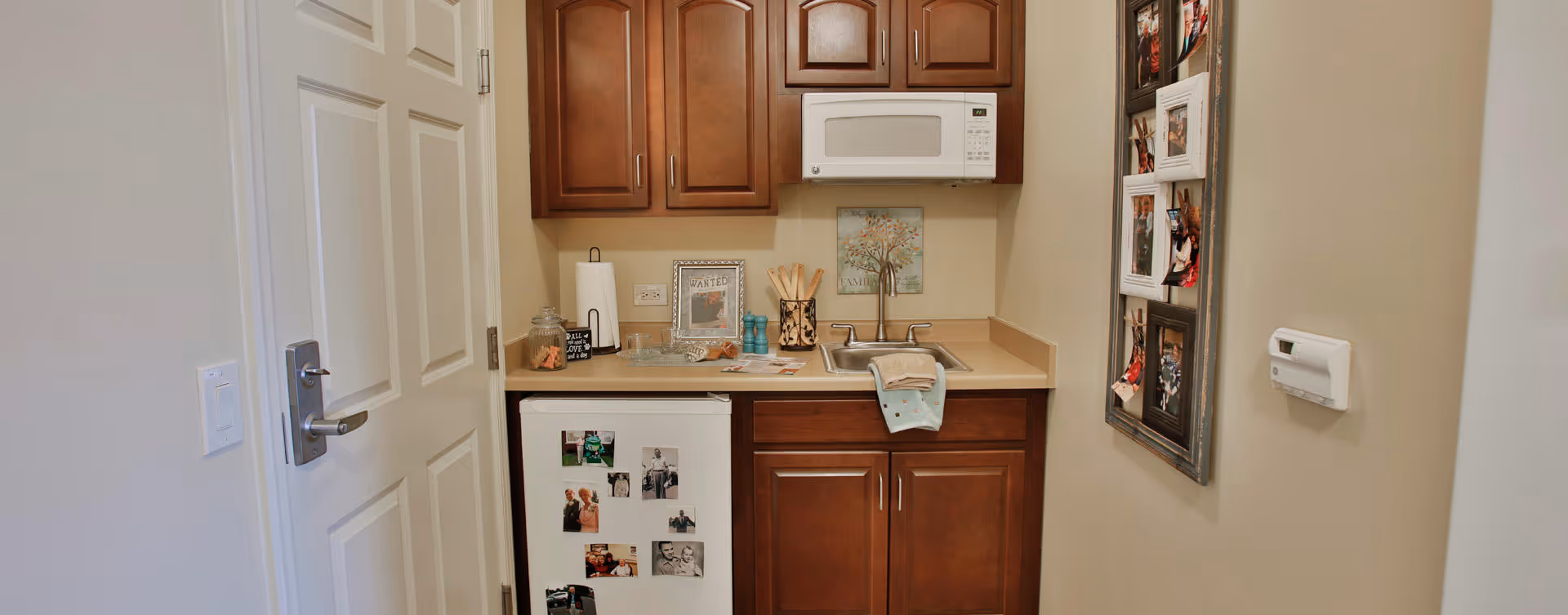 Small kitchenette area with wooden cabinets, a white microwave mounted above the counter, a small white refrigerator with photos attached, a sink with a towel hanging over the edge, and various decorative items on the countertop. A door is partially visible on the left, and a photo collage frame is mounted on the right wall.