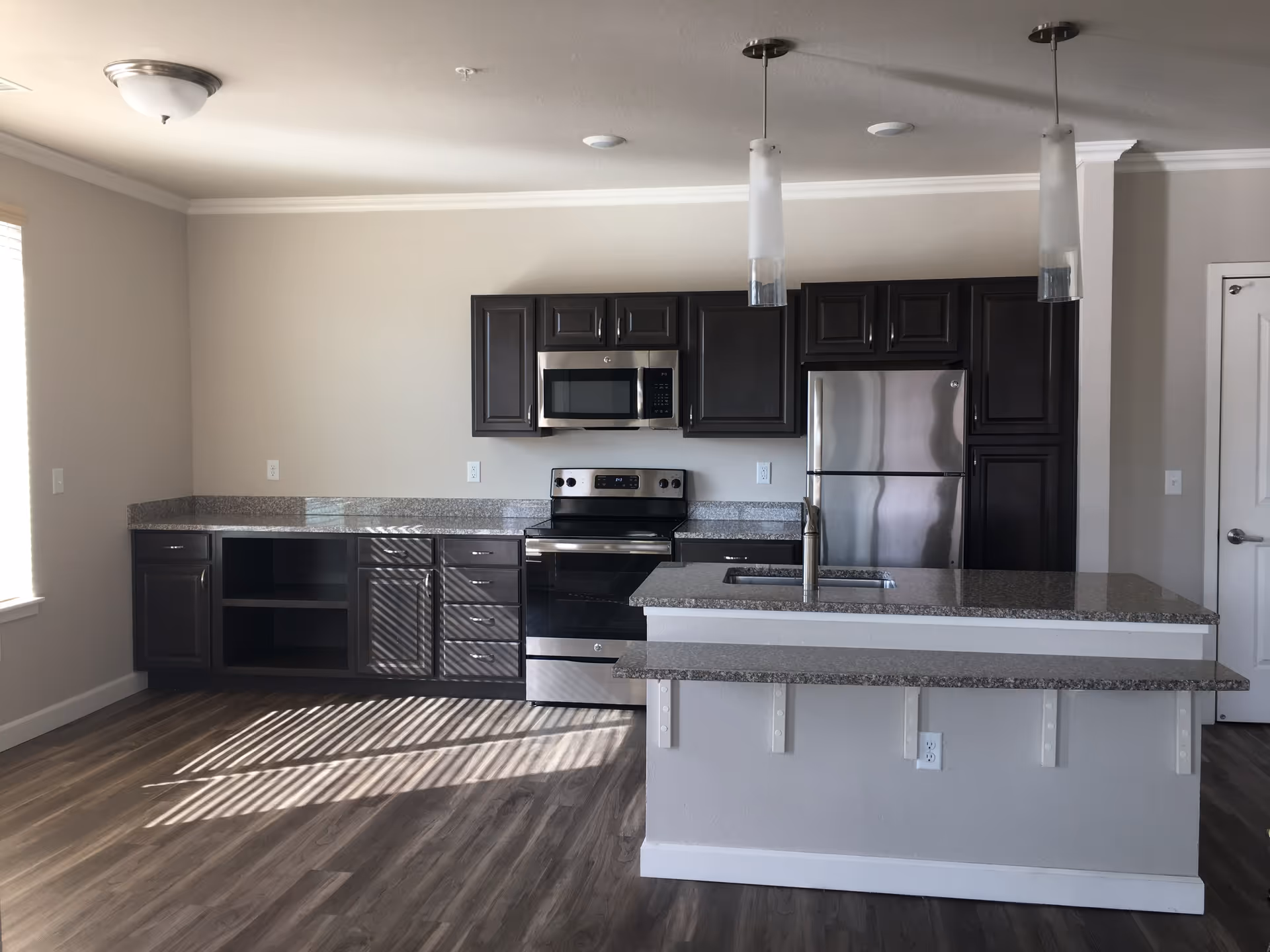 Modern kitchen with dark wood cabinets, stainless steel appliances including a refrigerator, microwave, and stove, granite countertops, a kitchen island with a sink, two pendant lights hanging from the ceiling, and wood flooring. Sunlight streams through a window casting shadows on the floor.