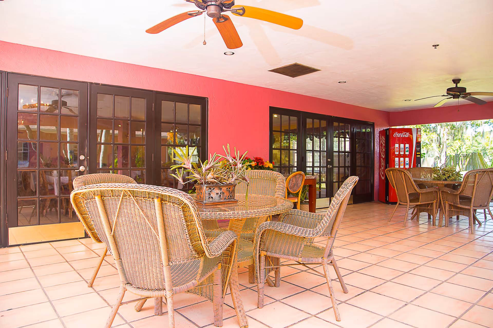 Covered outdoor patio area with wicker tables and chairs, ceiling fans, a red wall with large glass doors, and a Coca-Cola vending machine in the corner.
