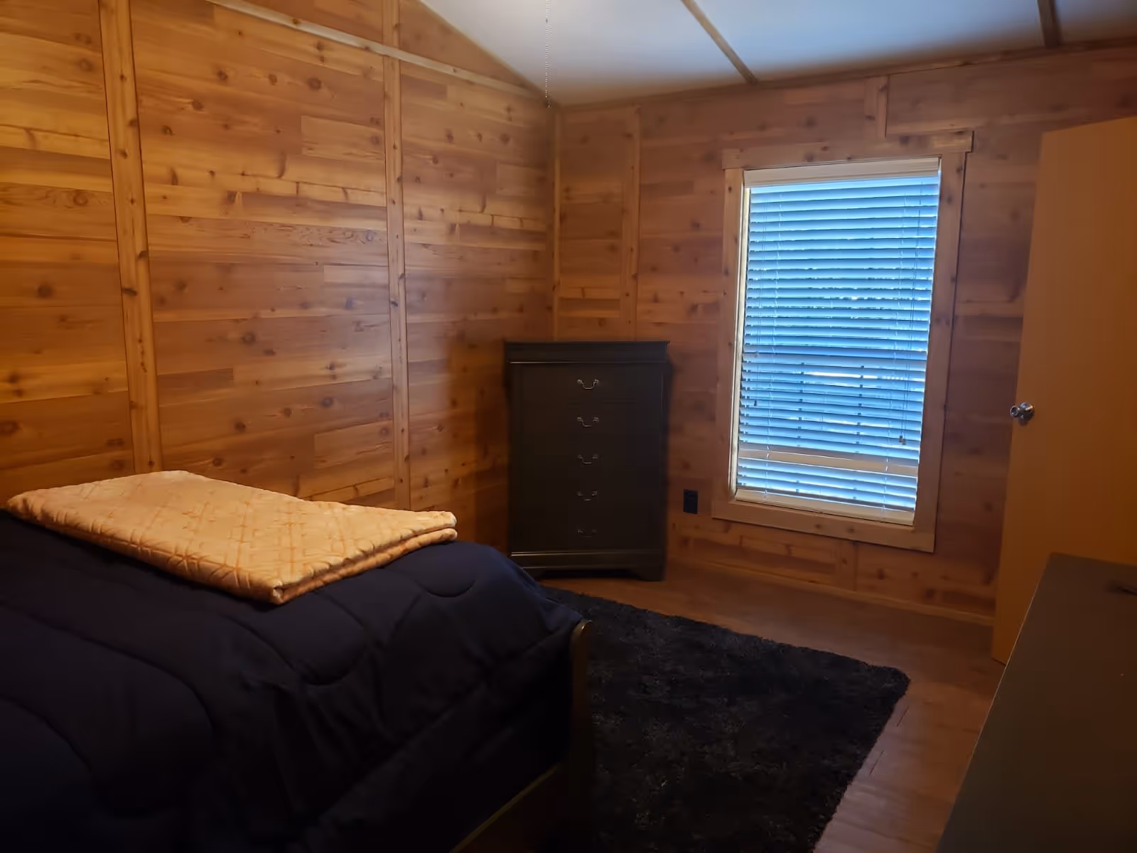 Small wood-paneled bedroom with a bed, folded blanket, dresser, window with blinds, and a door.