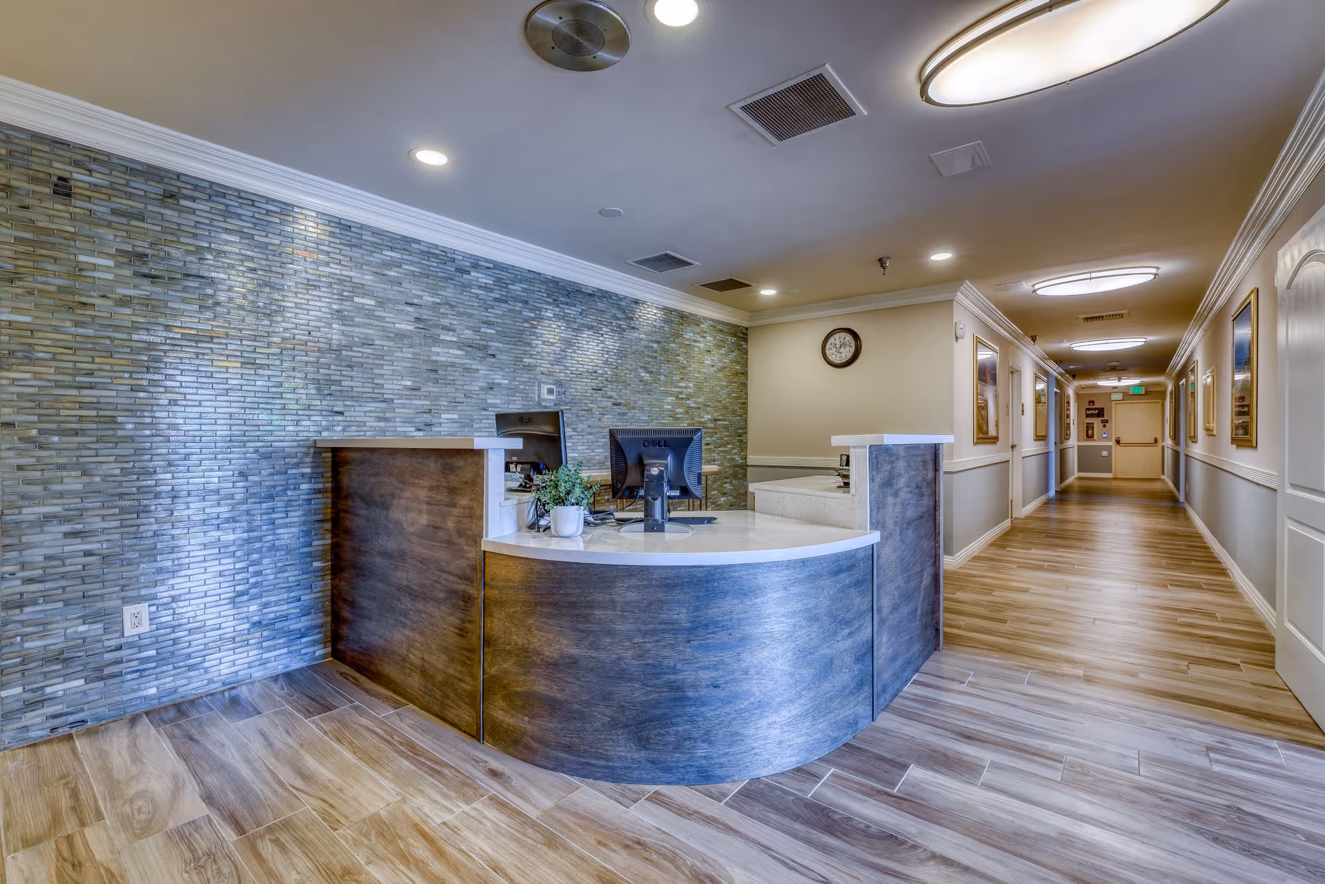 Reception desk area inside a senior living facility with a curved wooden desk, two computer monitors, a small potted plant, and a textured blue-gray tiled wall behind. The floor is wood-patterned tile, and a hallway with framed pictures and ceiling lights extends to the right.