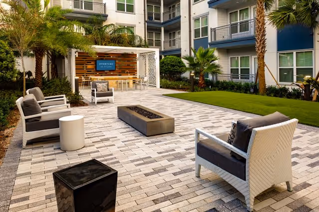 Outdoor patio area at Overture Dr. Phillips featuring modern seating with cushioned chairs around a rectangular fire pit, a pergola with a dining table and chairs, surrounded by palm trees and a multi-story residential building in the background.
