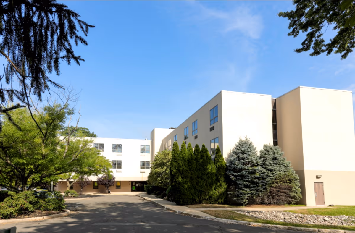 Front exterior of a three-story pale beige senior living building with landscaped trees and a driveway under a clear blue sky.