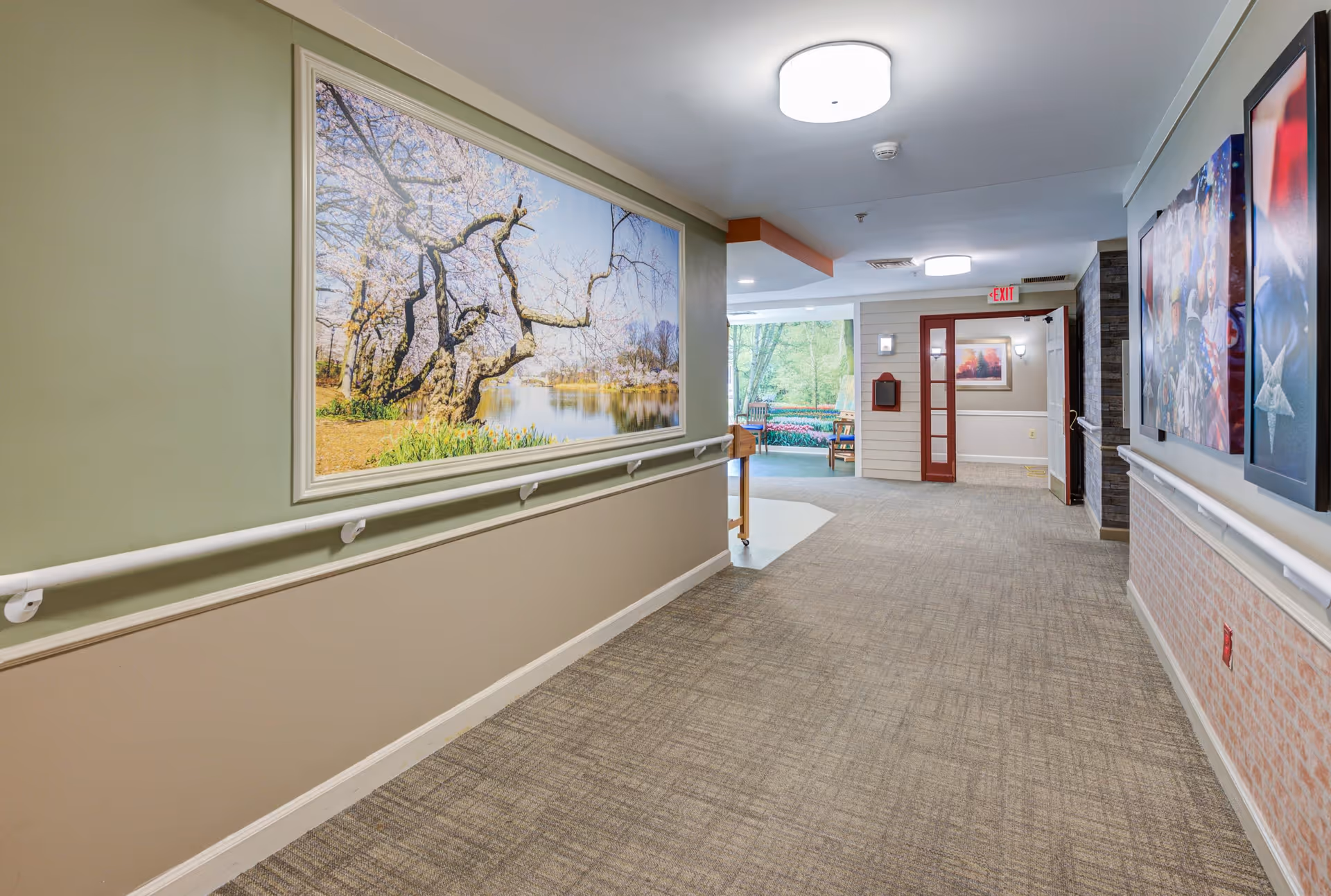 A well-lit hallway in a senior living facility with beige and green walls, carpeted floor, handrails on both sides, and large framed artwork depicting a tree by a lake on the left wall. The hallway leads to an open area with chairs and a large window showing greenery outside. There are additional framed pictures on the right wall and an exit sign above a door in the distance.