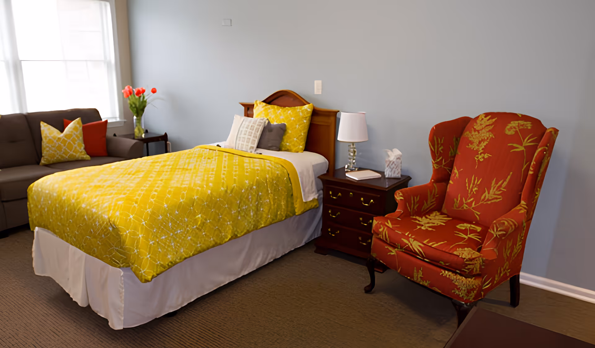 A neatly furnished bedroom with a bed covered in a yellow quilt, a red patterned armchair, a nightstand with a lamp, and a sofa by the window.