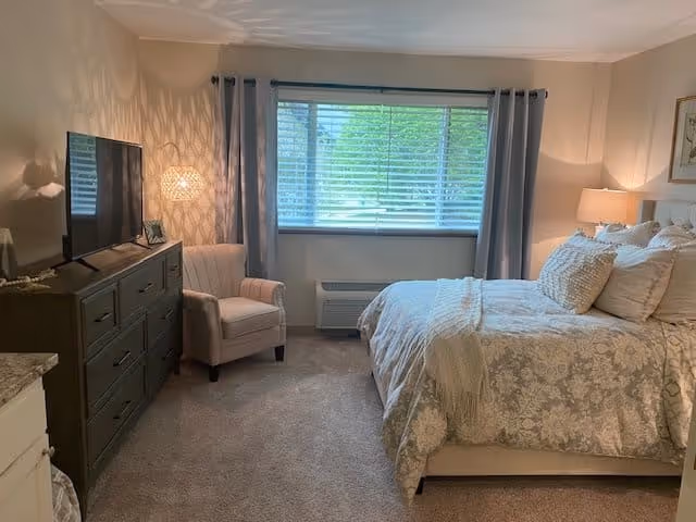 A cozy bedroom with a large window covered by blinds and gray curtains. The room features a bed with floral-patterned bedding and multiple pillows, a bedside table with a lamp, a cushioned armchair, and a dark dresser with a flat-screen TV on top. The walls are light-colored, and the carpet is beige.