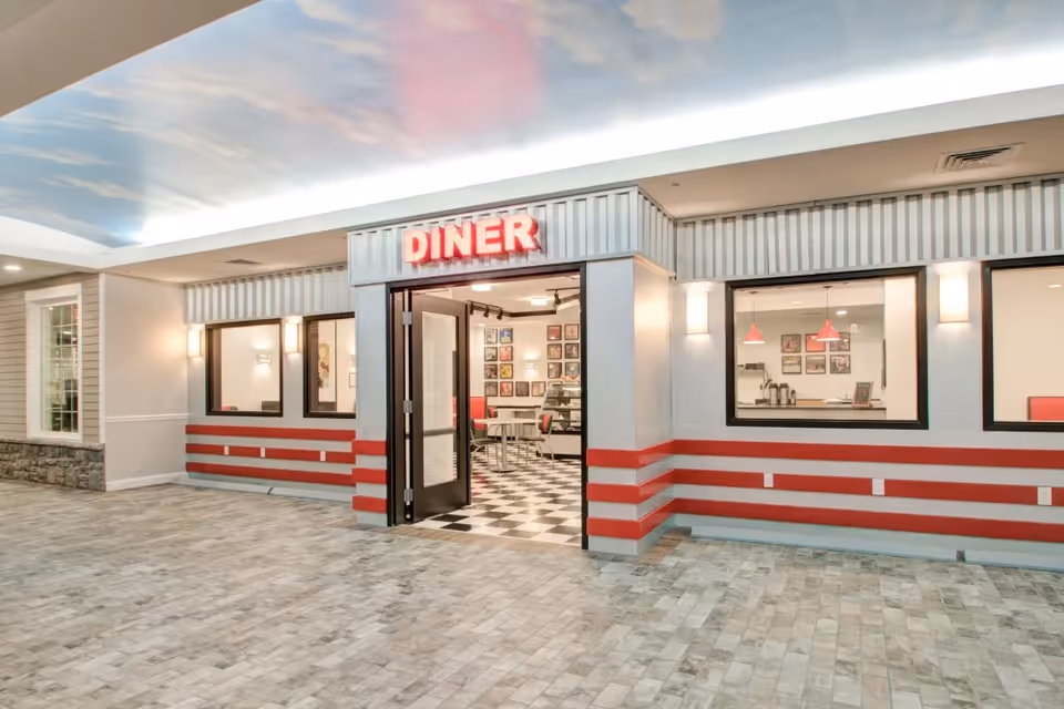 Entrance to a retro-style diner inside a facility, with a red neon sign reading 'DINER' above the open double doors. The exterior walls have red horizontal stripes, and the floor is tiled with a stone pattern. Inside, black and white checkered flooring and red chairs are visible, along with framed pictures on the walls. The ceiling outside is painted with a sky and clouds motif.