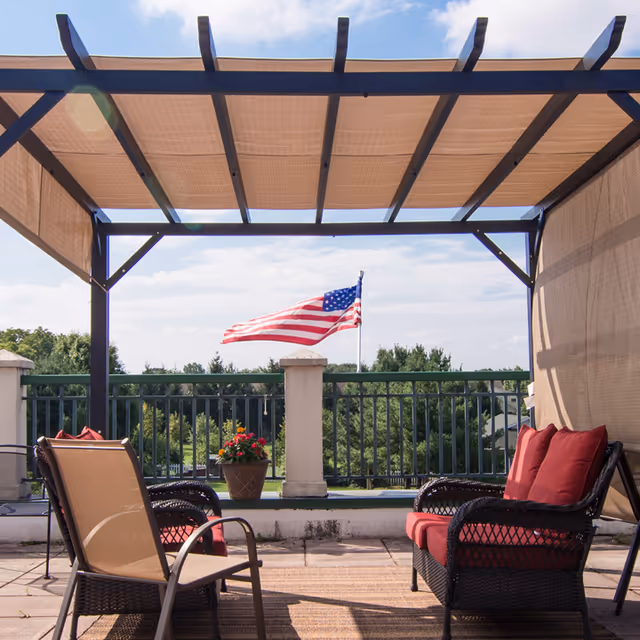 Outdoor seating area with a pergola overhead, featuring two wicker chairs with red cushions and a beige chair around a small table. An American flag is visible in the background, along with greenery and a partly cloudy sky.