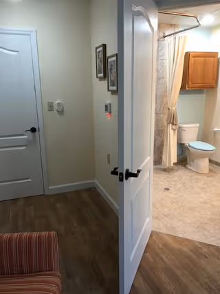 View of an interior space showing a partially open white door leading to a bathroom with a toilet, a wooden cabinet, and a shower curtain. The foreground includes a corner of a striped upholstered chair and a hallway with two framed pictures on the wall.