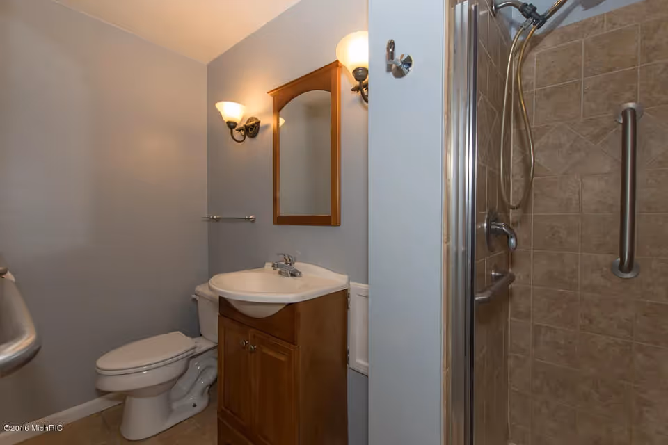 Small bathroom with a toilet, wooden vanity and mirror, wall sconces, and a tiled shower with grab bar.