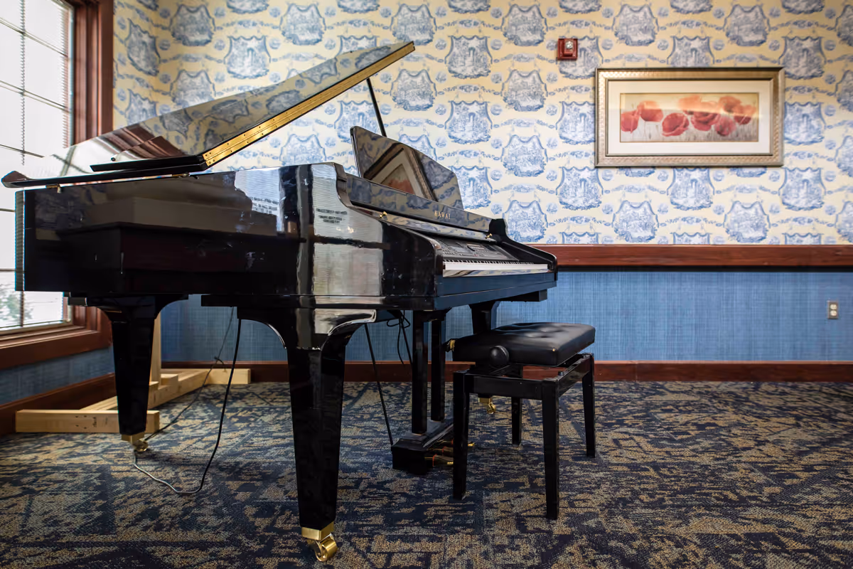 A black grand piano and bench sit on patterned carpet in a decorated interior room with floral wallpaper and framed artwork.