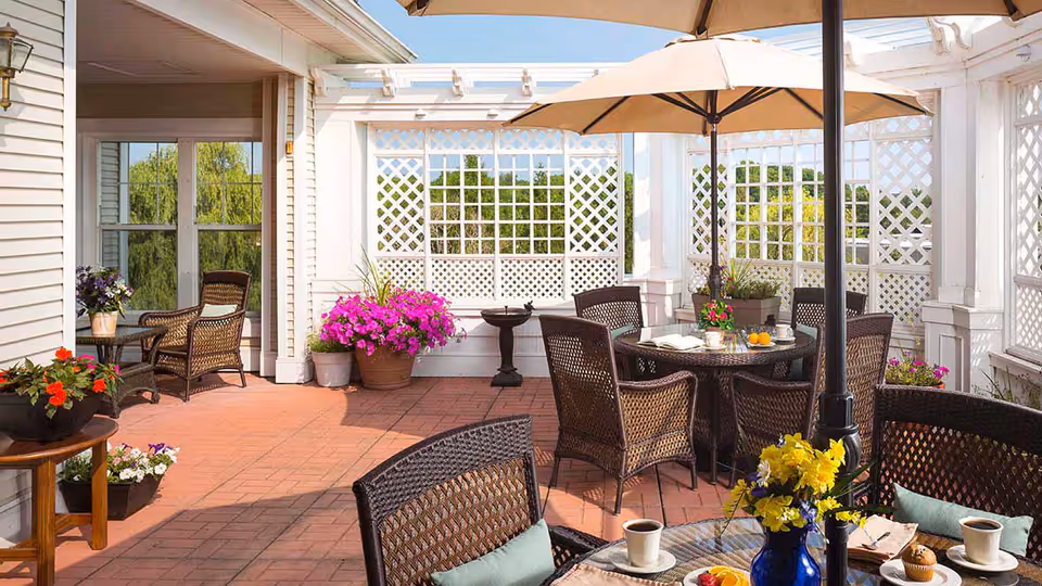 Outdoor patio area with wicker chairs and tables under large umbrellas. The space is decorated with colorful potted flowers and surrounded by white lattice fencing. The patio floor is made of red tiles, and there is a birdbath in the background.